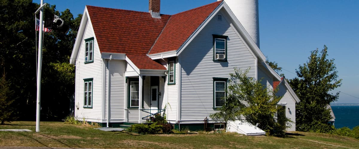 A white lighthouse with a red roof, an American flag, and a view of the ocean.