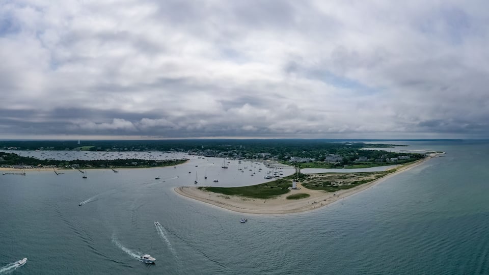 An aerial view of a coastal area with a sandy beach, a small island with buildings, and numerous boats in the water.