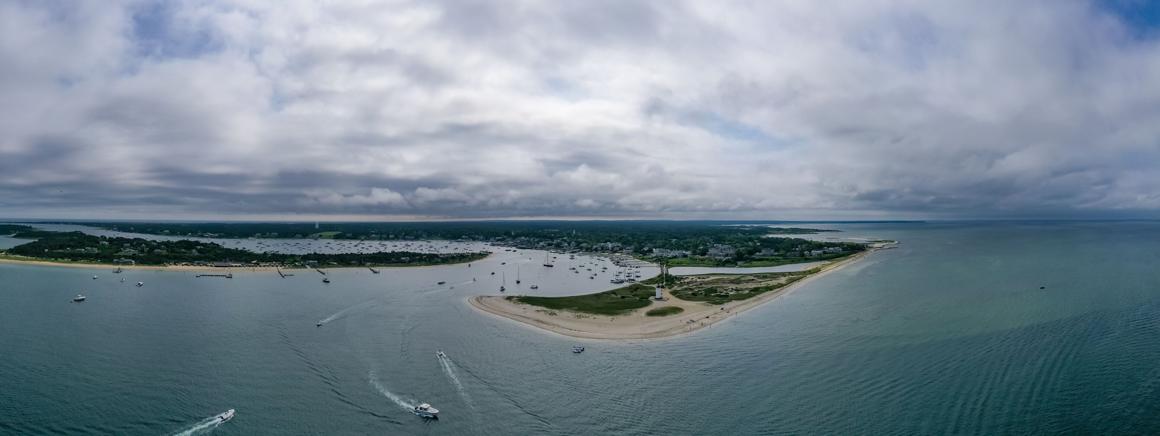 An aerial view of a coastal area with a sandy beach, a small island with buildings, and numerous boats in the water.