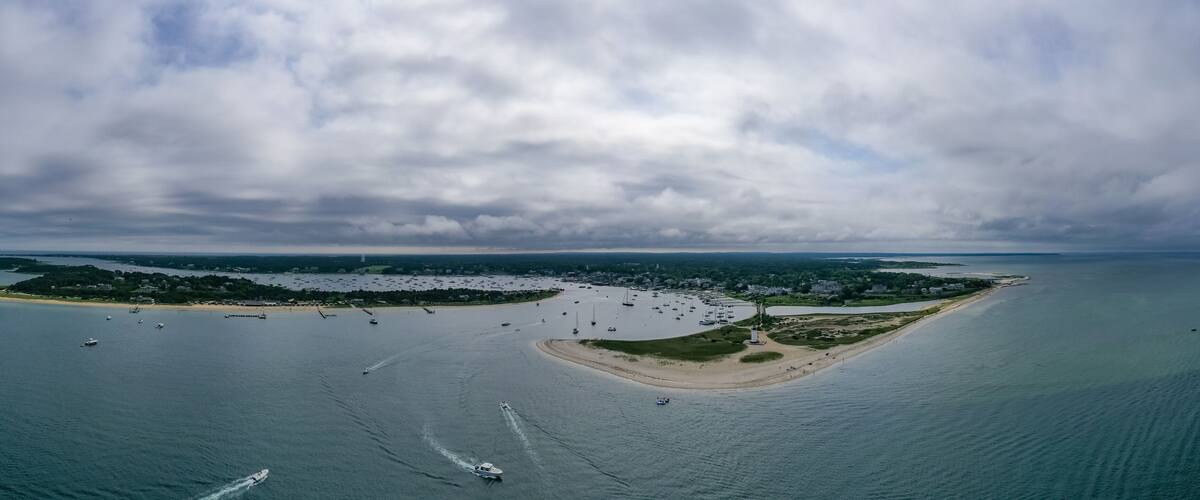 An aerial view of a coastal area with a sandy beach, a small island with buildings, and numerous boats in the water.