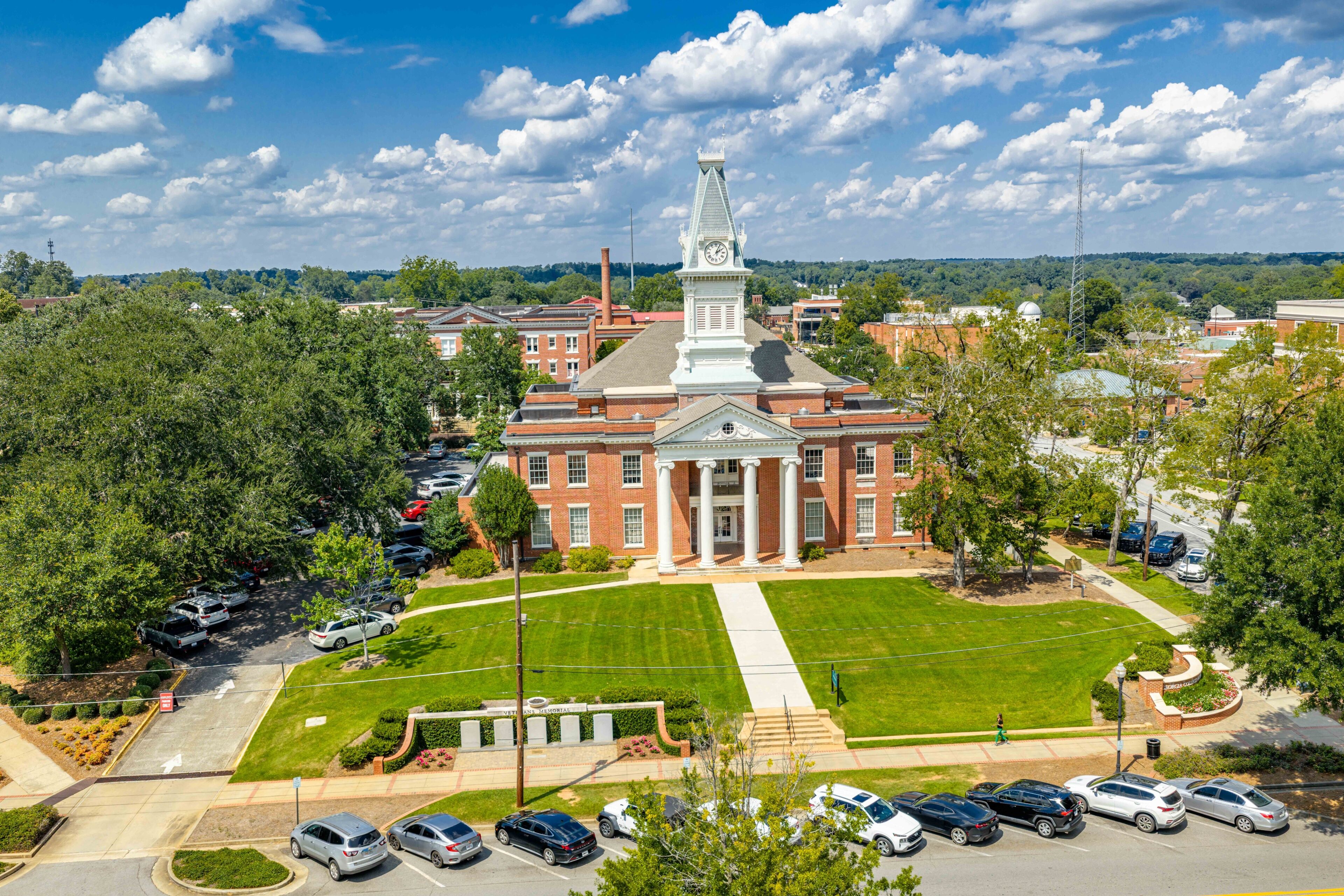A large red-brick building with a central tower and columns, surrounded by a well-maintained lawn and parked cars.