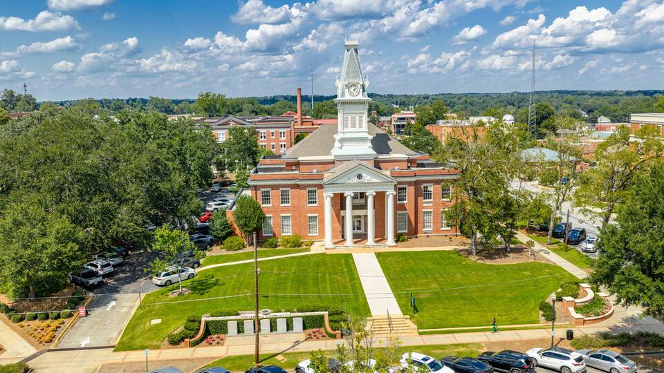 A large red-brick building with a central tower and columns, surrounded by a well-maintained lawn and parked cars.