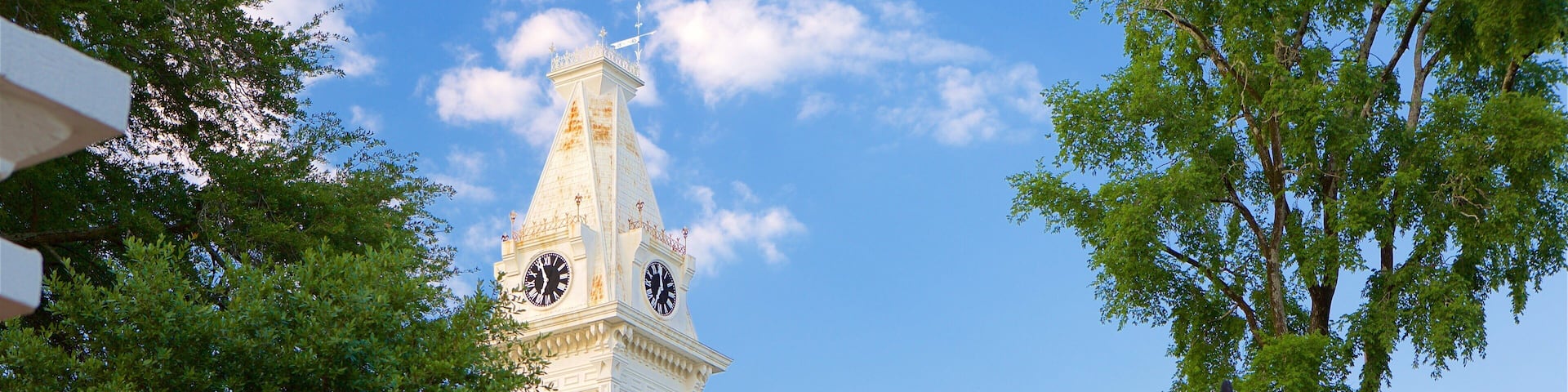 A white clock tower with a dome and a steeple, surrounded by trees and a building with a red brick facade.