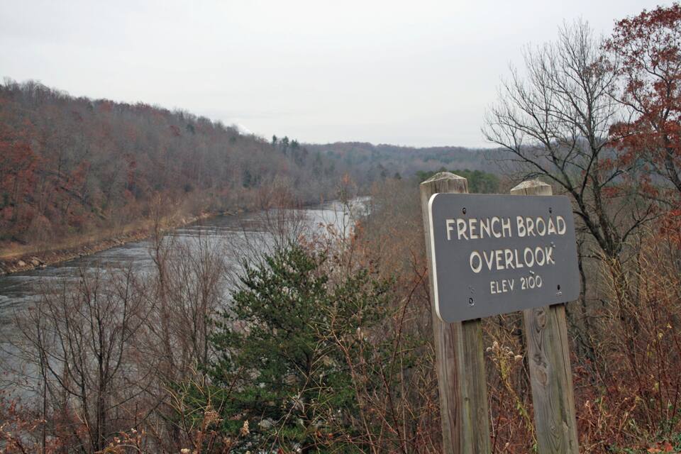 A sign for French Broad Overlook with an elevation of 2100, surrounded by autumn foliage and a river.