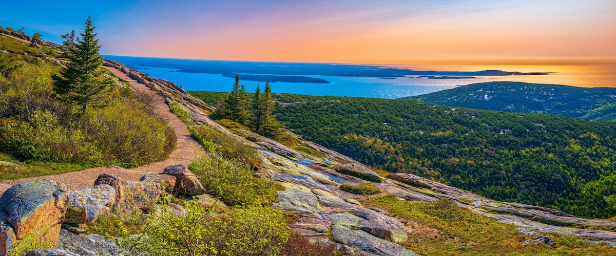 A rocky hiking trail with dense forest and a view of a large body of water during sunset.