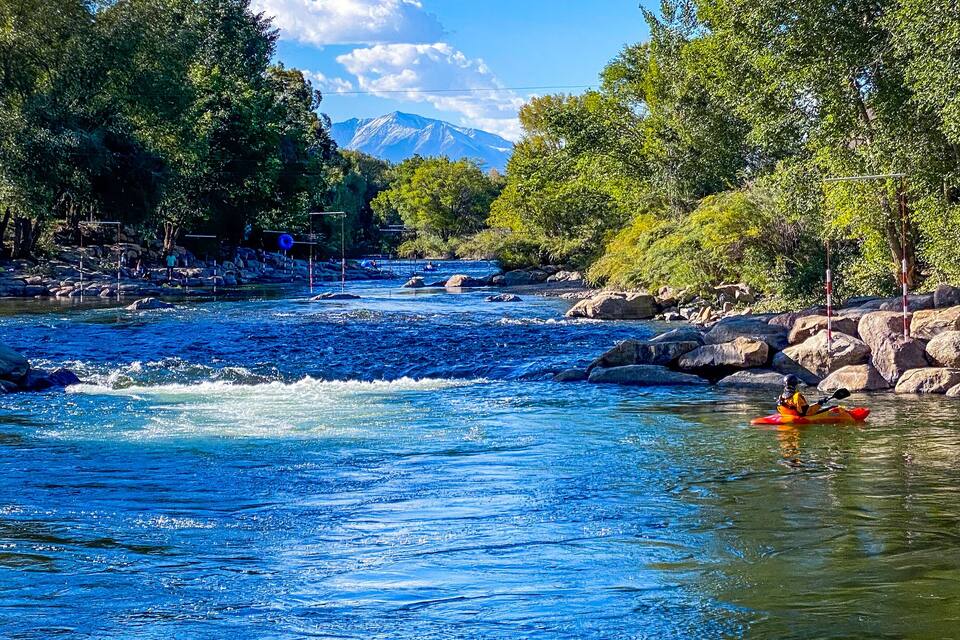 A river with a kayaker, surrounded by trees and rocks, with a mountain in the background.