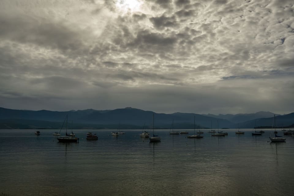 Um lago calmo com vários barcos à vela ancorados perto da margem, rodeado por montanhas sob um céu nublado.