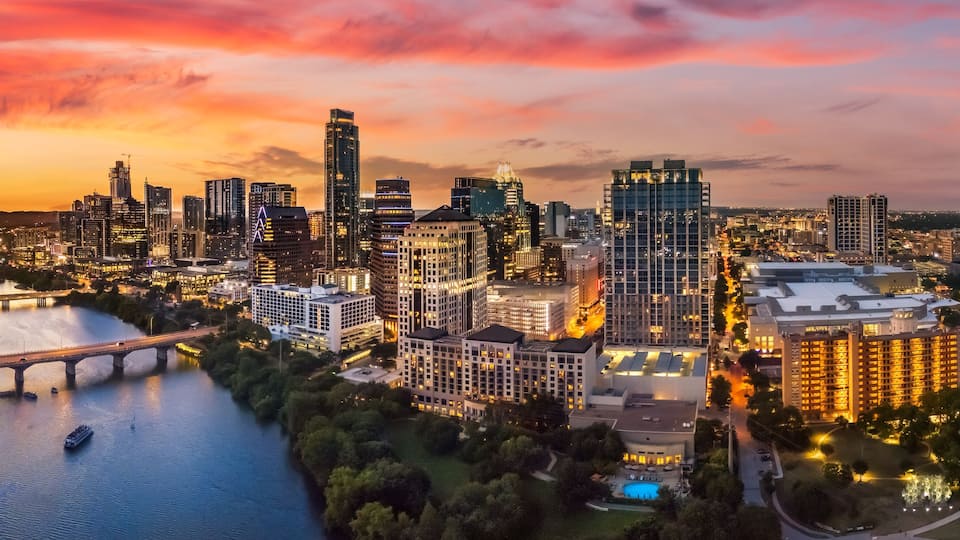 A cityscape at sunset with a river, a bridge, and modern high-rise buildings.