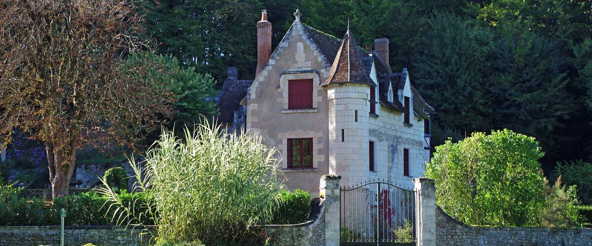 A stone house with a red window and a black gate, surrounded by lush greenery and trees.