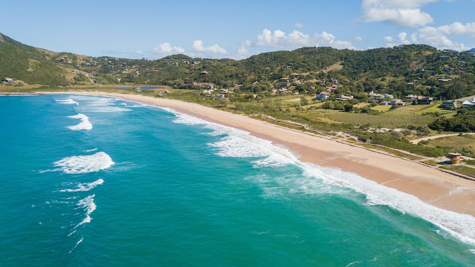 A coastal landscape with a turquoise beach, a clear sky, and a hilly background with scattered houses.