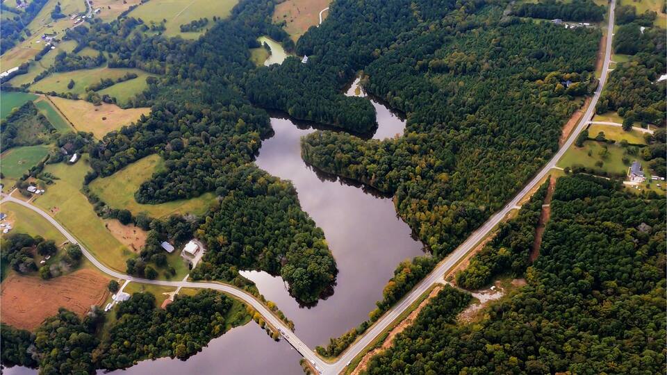 I fly over a very serene scene of a lake with a bridge, the sun glints off the lake and the clouds can be seen in the reflection off the lake. Very calming and a great peaceful shot. Randleman Lake in