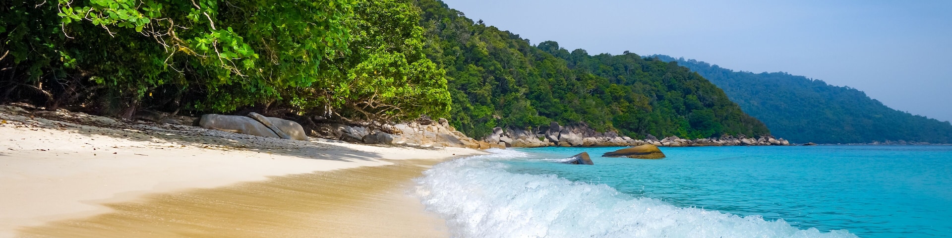 A sandy beach with waves, a forested hillside, and a clear blue sky.