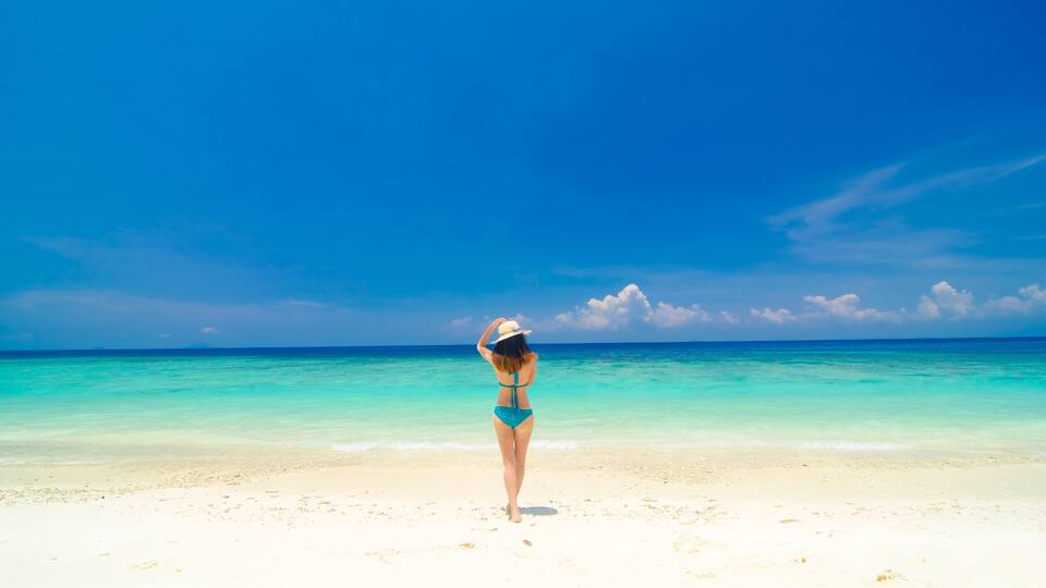 A woman in a blue bikini and white hat standing on a sandy beach with clear turquoise water and a blue sky.