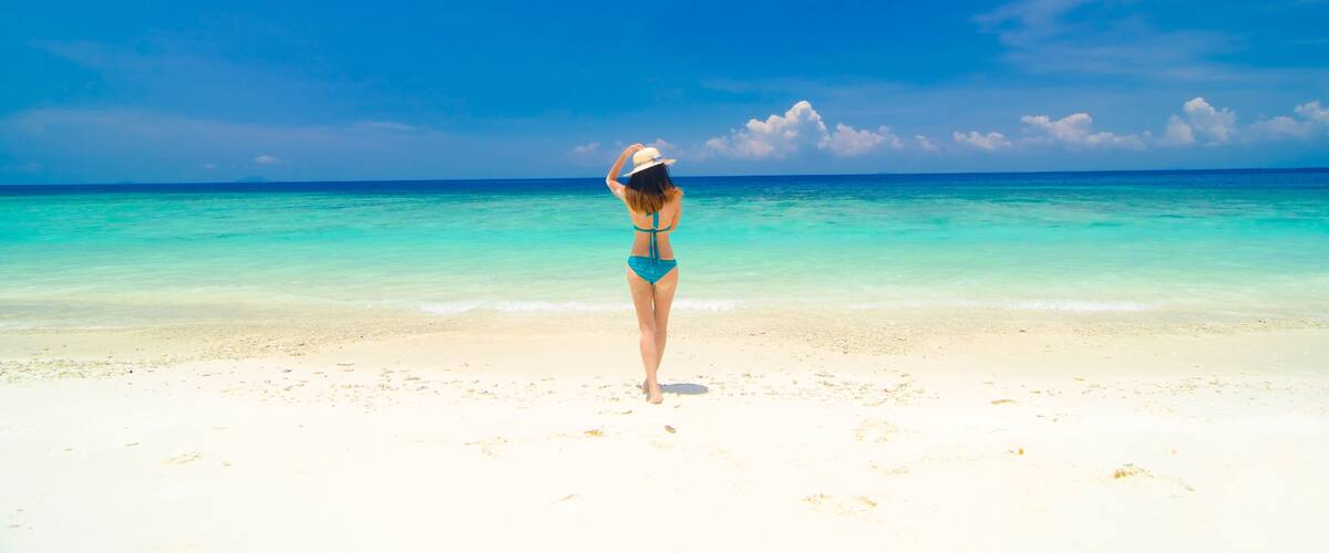 A woman in a blue bikini and white hat standing on a sandy beach with clear turquoise water and a blue sky.