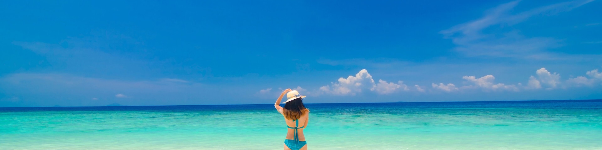 A woman in a blue bikini and white hat standing on a sandy beach with clear turquoise water and a blue sky.