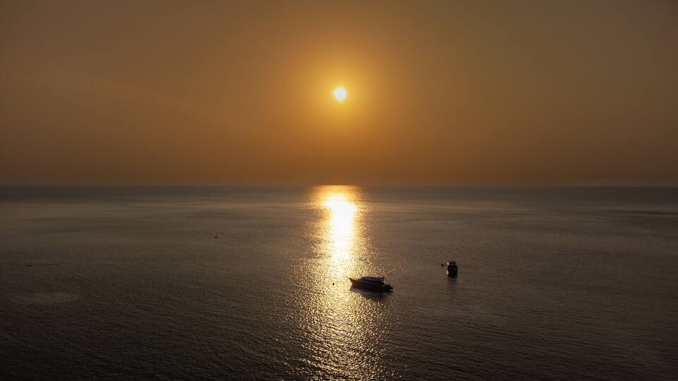 A sunset over a calm sea with a boat and a person standing in the water.