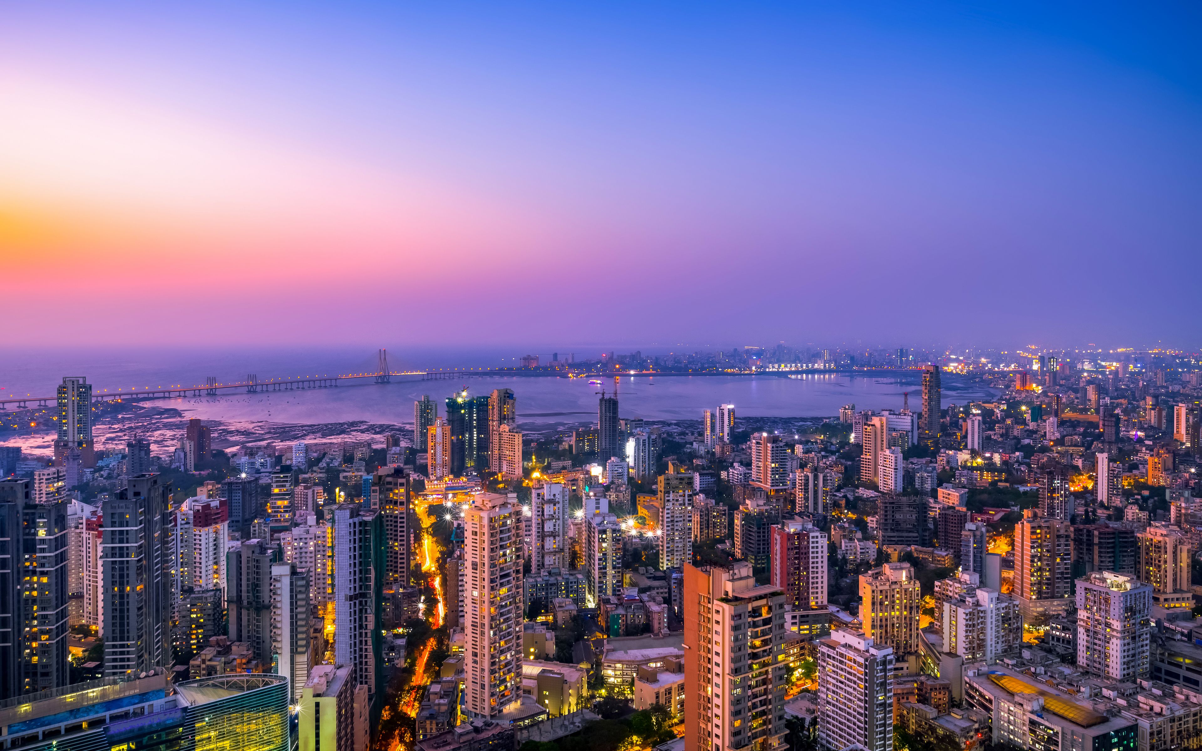 A cityscape at dusk with illuminated buildings and a bridge in the distance.