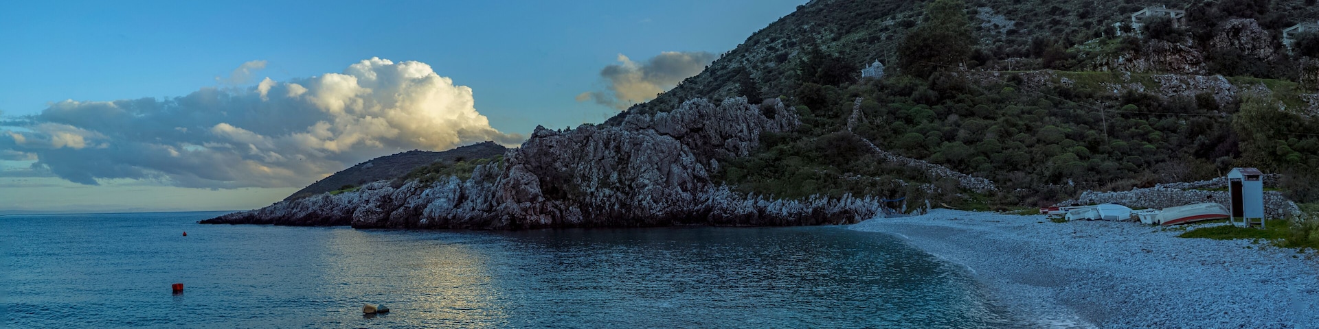 A pebble beach with clear blue water, a rocky hill, and a small building.