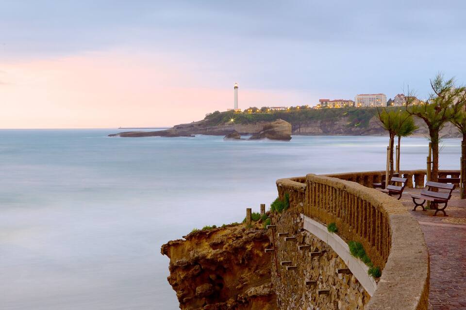 A coastal scene with a stone wall, benches, and a lighthouse in the background.