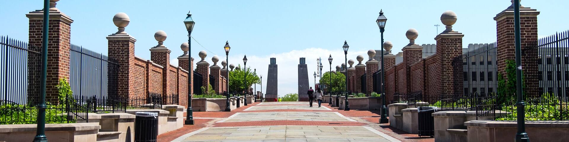 A paved pathway flanked by brick walls and street lamps.
