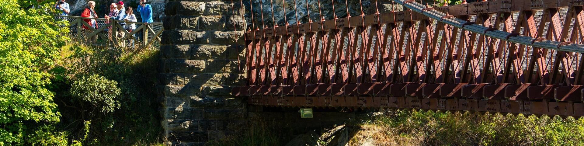 A suspension bridge with stone pillars and a metal deck, surrounded by rocky cliffs and greenery.