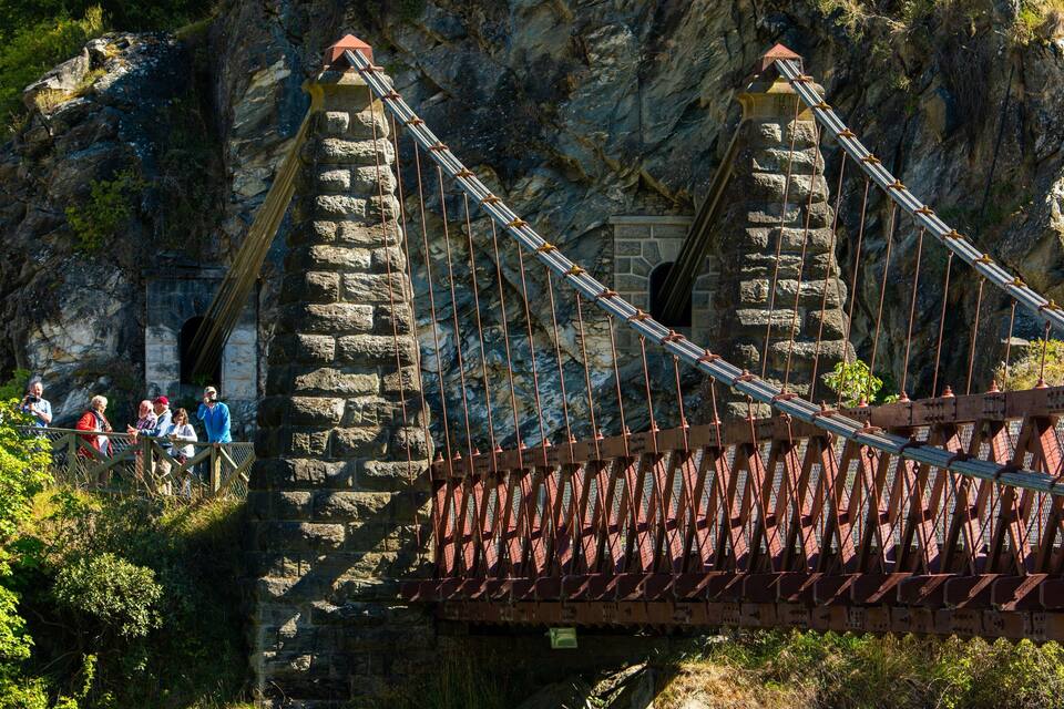 Un puente colgante con pilares de piedra y plataforma metálica, rodeado de riscos y vegetación.