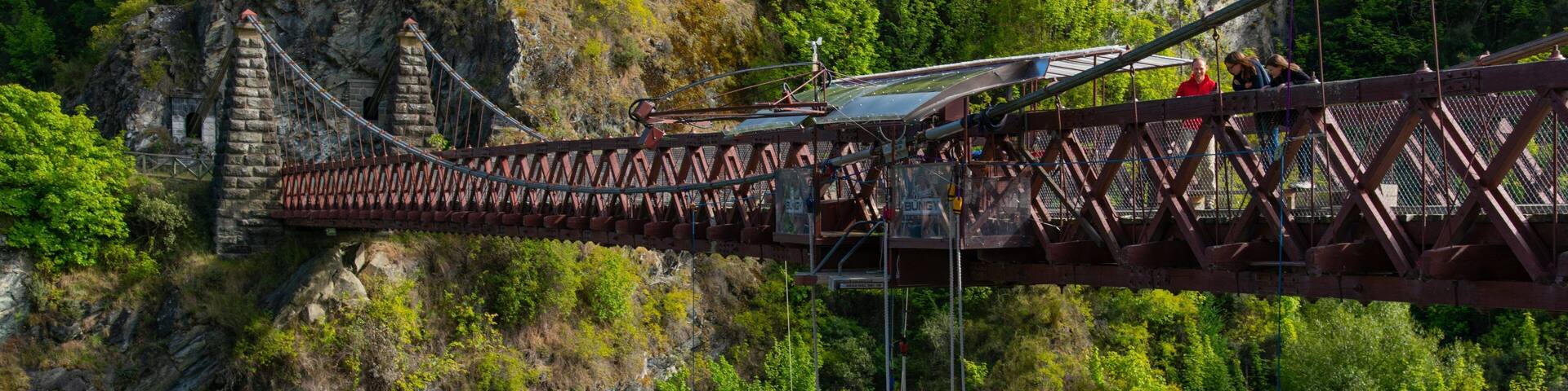 A suspension bridge with a stone tower, surrounded by lush greenery and rocky cliffs.