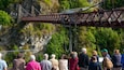 A suspension bridge with a stone tower, surrounded by lush greenery and rocky cliffs.