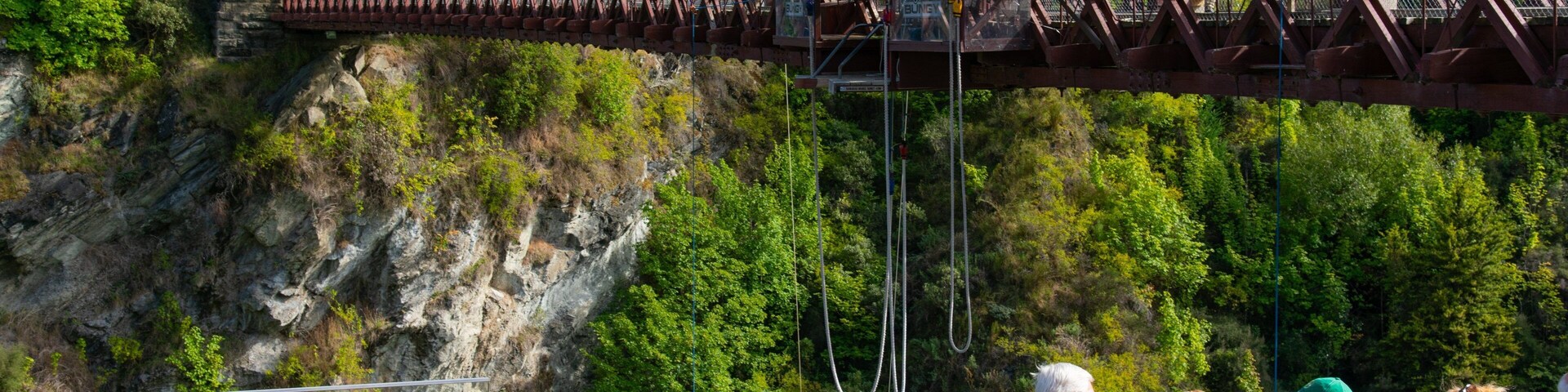 Uma ponte suspensa com uma torre de pedra, rodeada por vegetação exuberante e penhascos rochosos.