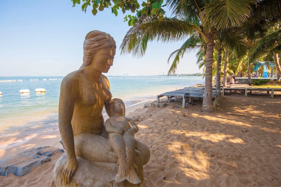 En statue af en kvinde, der holder et barn, på en sandstrand med palmer og udsigt til havet.