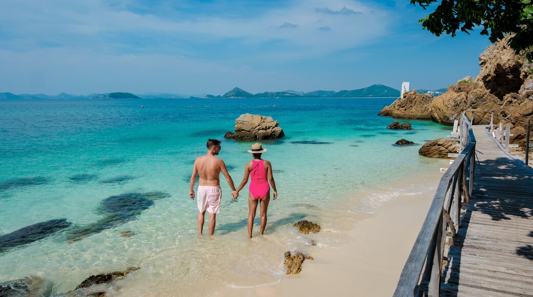A couple walking on a wooden pier towards a clear turquoise sea with rocky outcrops and distant mountains.