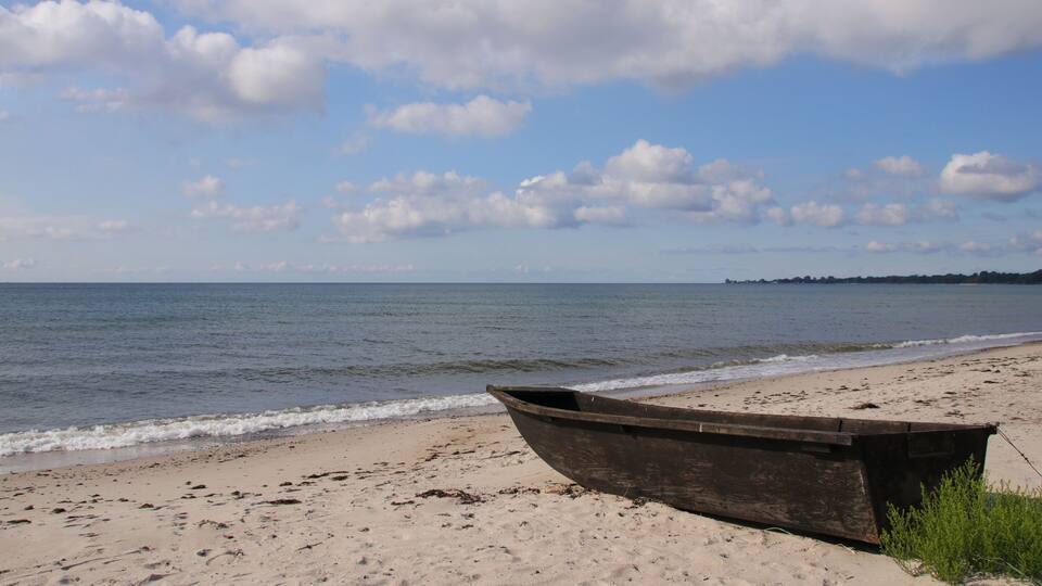A wooden boat on a sandy beach with a calm sea and a partly cloudy sky.