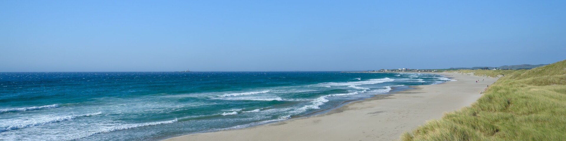 Una playa con olas suaves, cielo despejado y dunas cubiertas de hierba.
