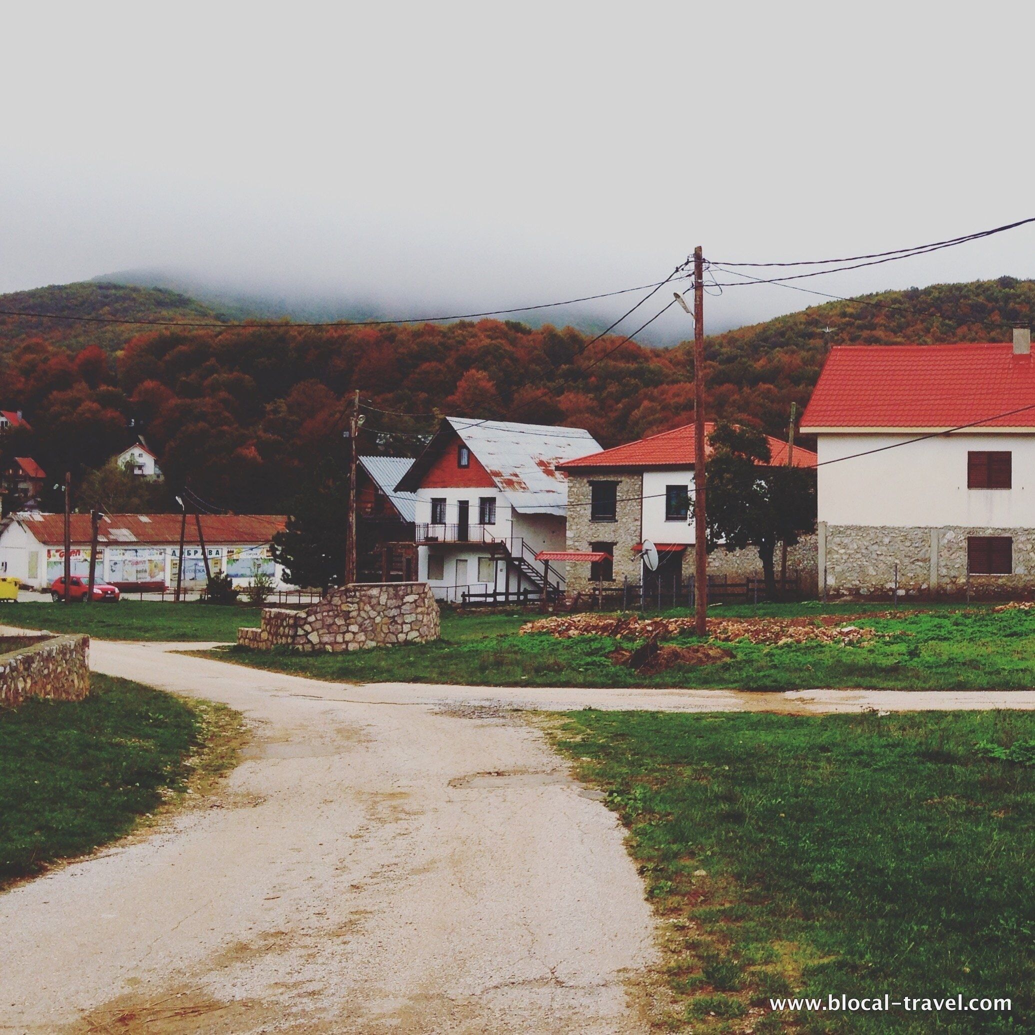 A rural road with houses, a stone wall, and autumn-colored trees in the background.
