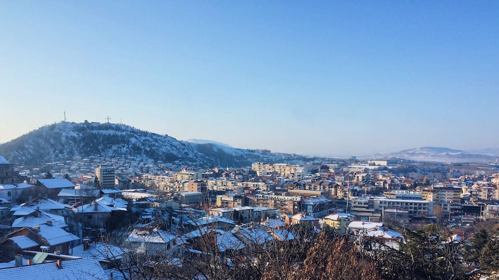 A snow-covered cityscape with buildings and a hill in the background.