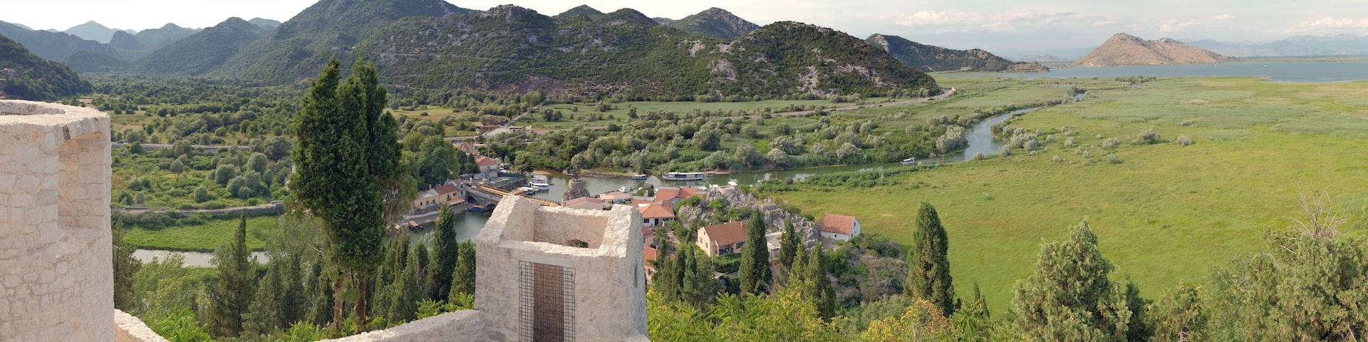 A historic stone tower overlooking a river, surrounded by lush greenery and mountains.