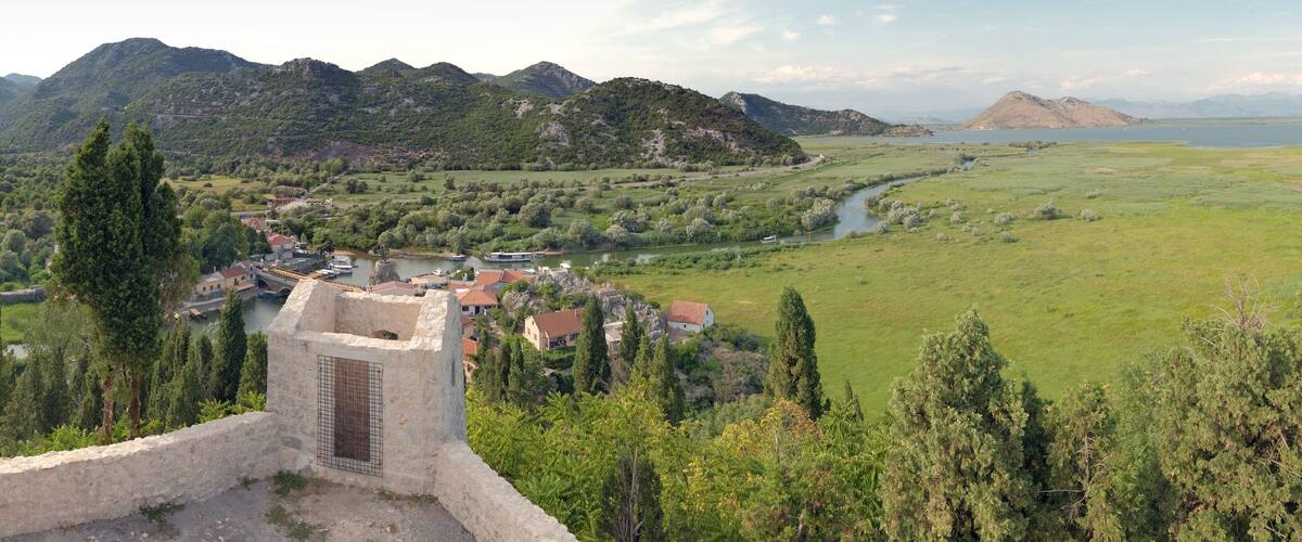 A historic stone tower overlooking a river, surrounded by lush greenery and mountains.