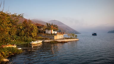Un lago sereno con una pequeña casa sobre un muelle de piedra, rodeado de exuberante vegetación y una montaña al fondo.