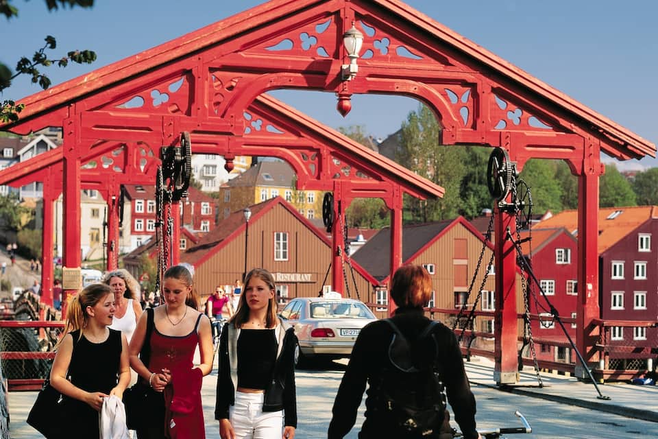 A red metal bridge with intricate designs, surrounded by buildings and greenery.