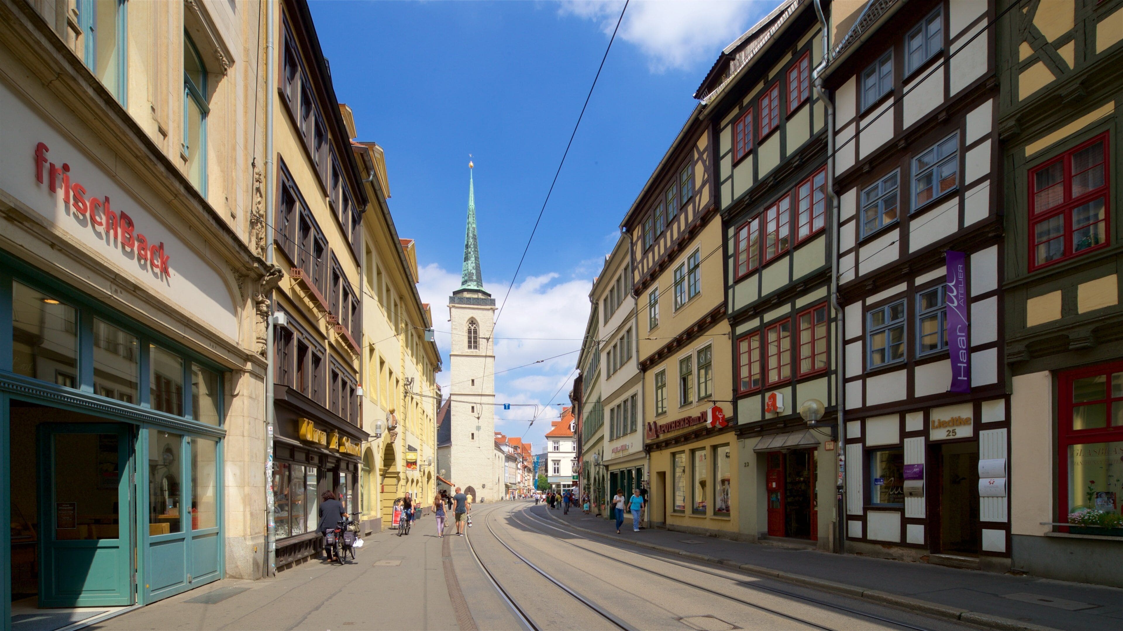A street with historic buildings, a church spire, and tram tracks.
