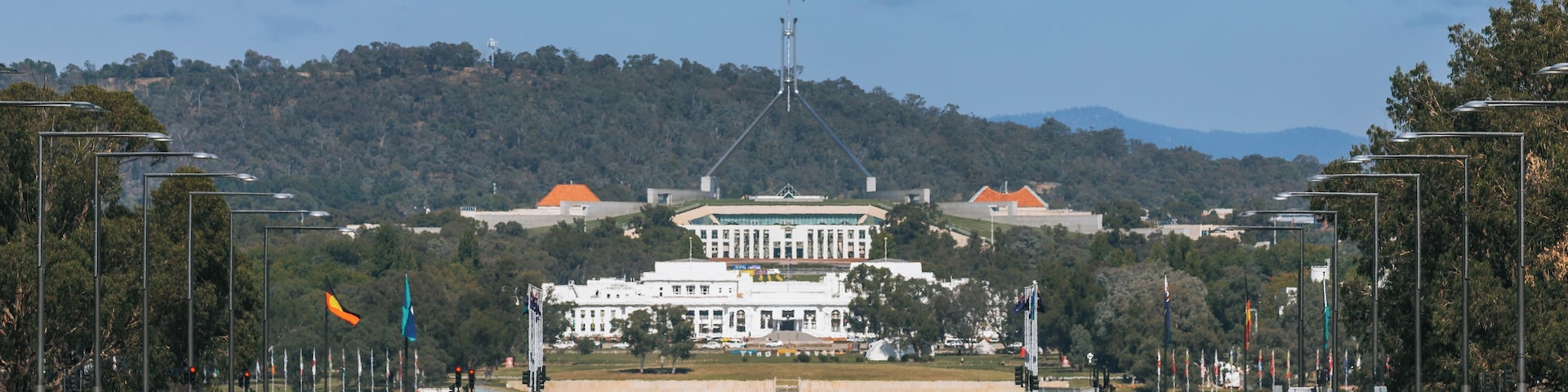A large white building with a flagpole in front, surrounded by trees and hills in the background.