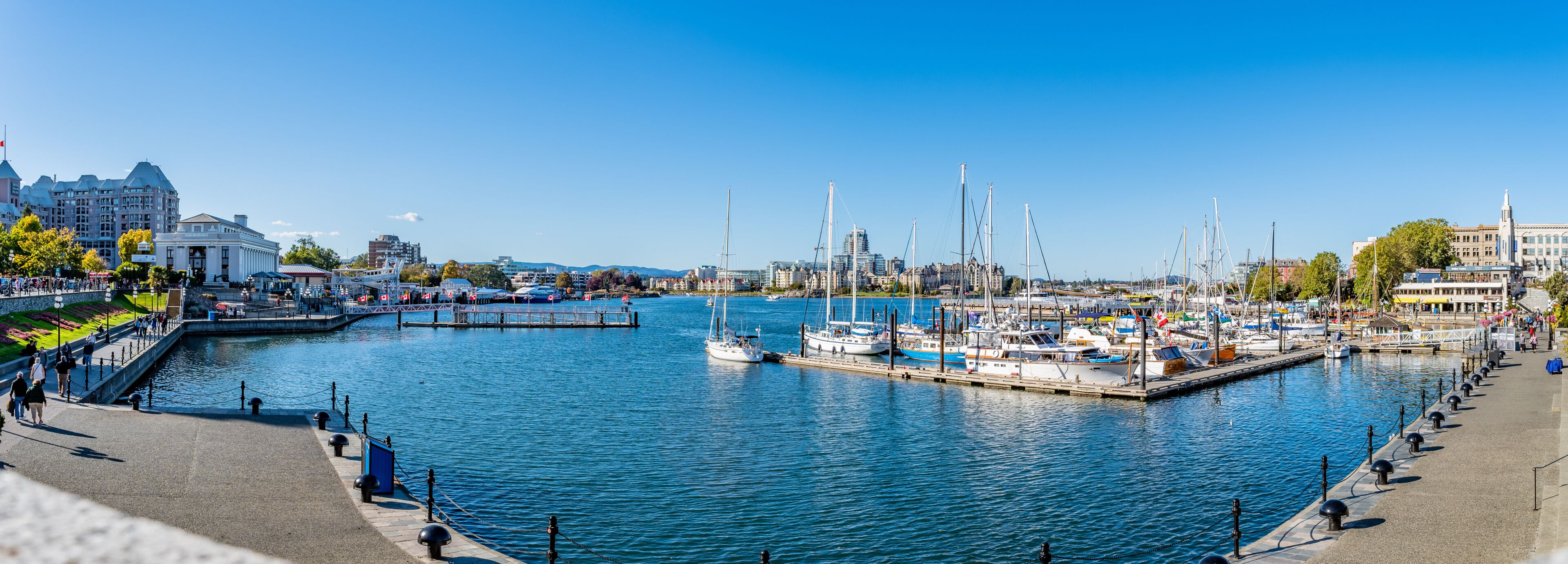 A marina with boats docked, a waterfront promenade, and buildings in the background.
