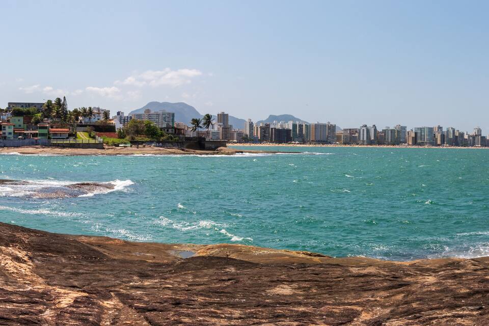 A coastal cityscape with a rocky shoreline, a beach, and a densely built urban area with high-rise buildings.
