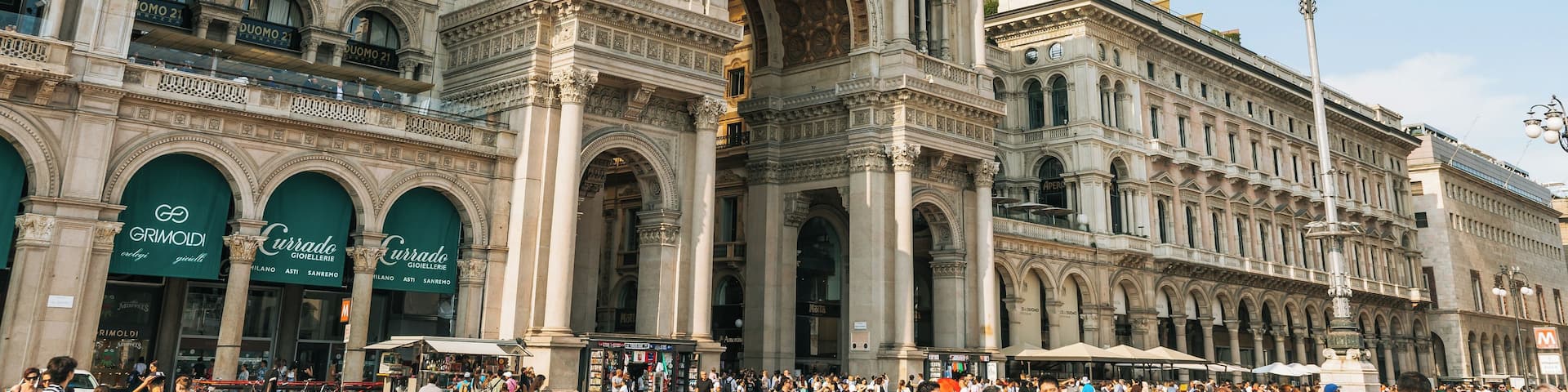 A historic building with ornate architecture, including columns and arches, and a bustling square with people and shops.
