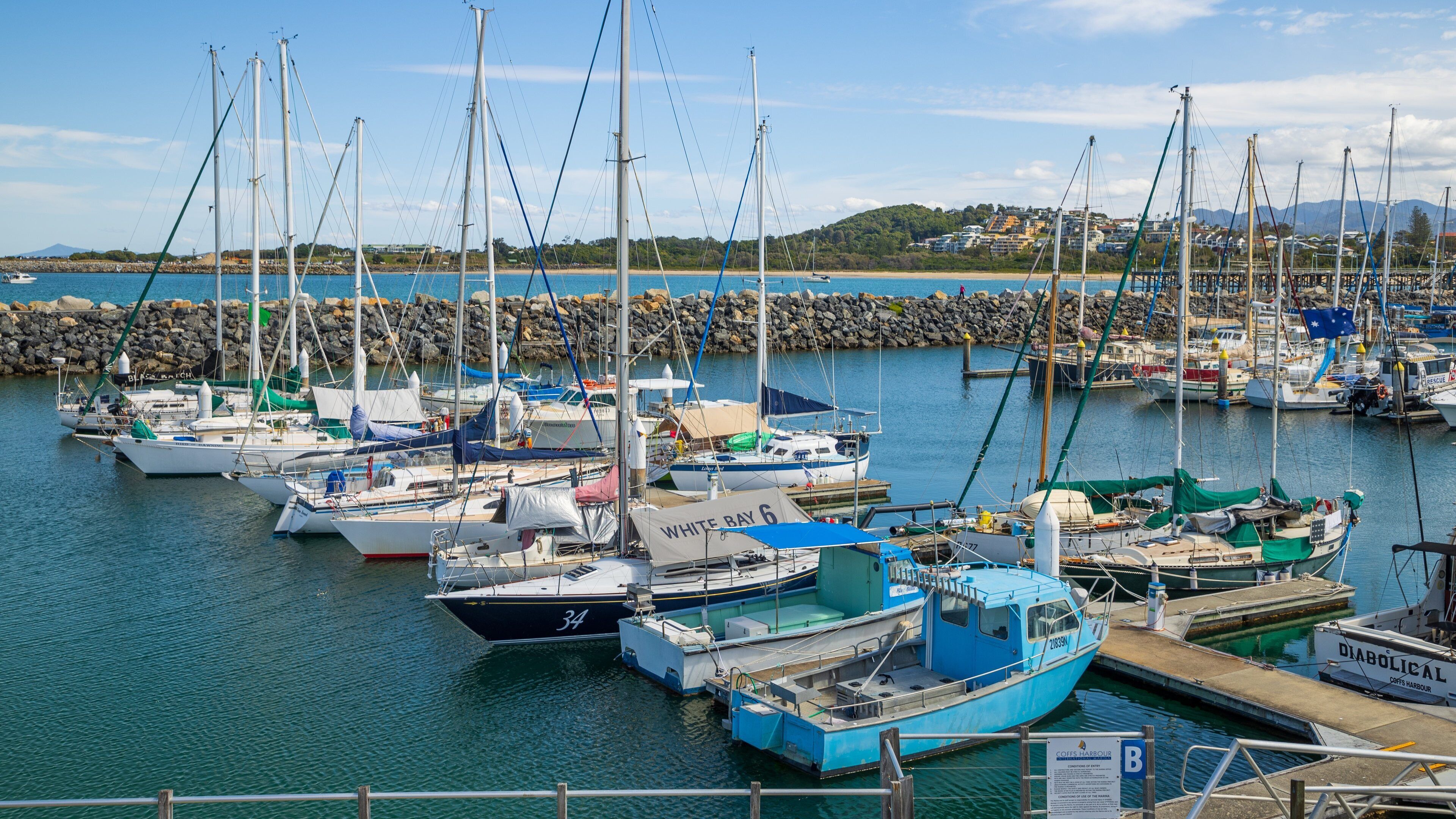 A marina with various sailboats docked, a rocky breakwater, and a distant mountain range.