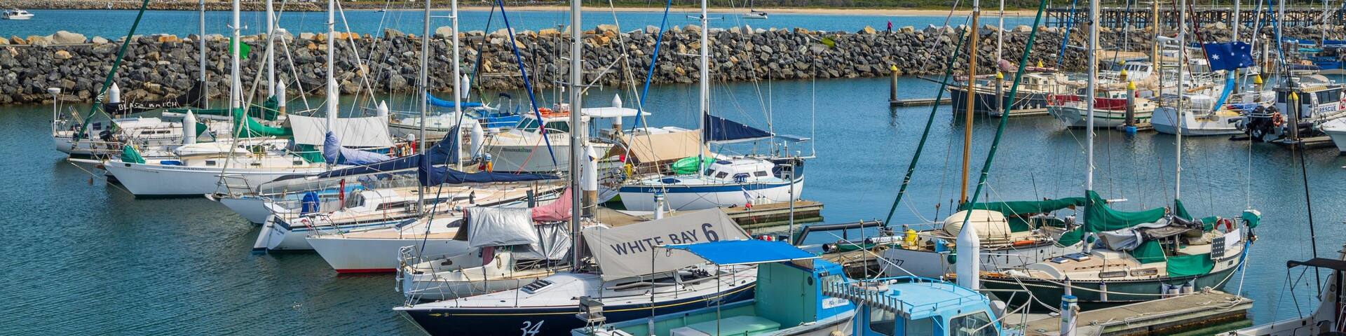 A marina with various sailboats docked, a rocky breakwater, and a distant mountain range.