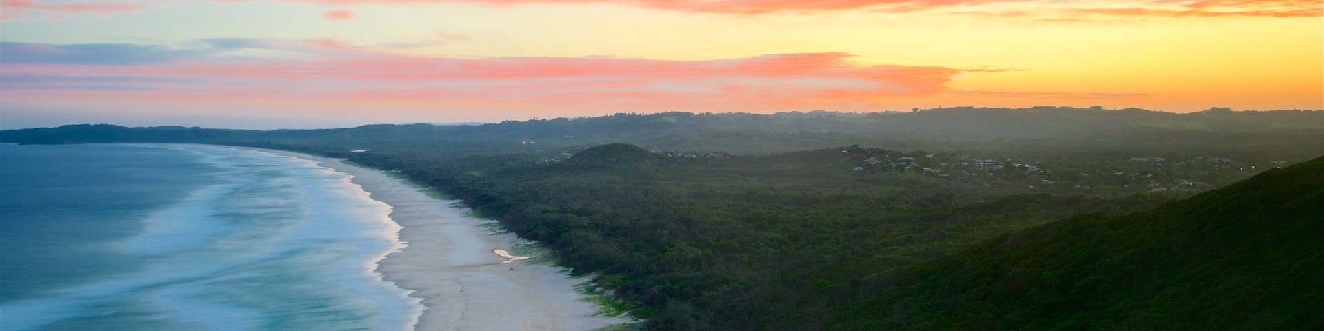 Paisagem costeira com praia, colinas arborizadas e pôr do sol.
