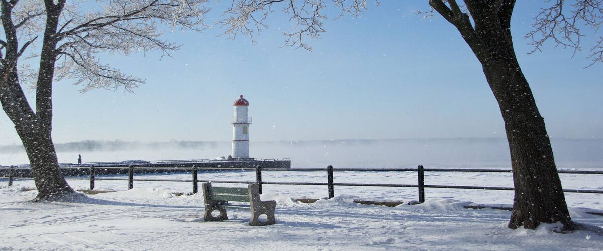 A snow-covered park with a bench, trees, a lighthouse, and a railing along a waterfront.