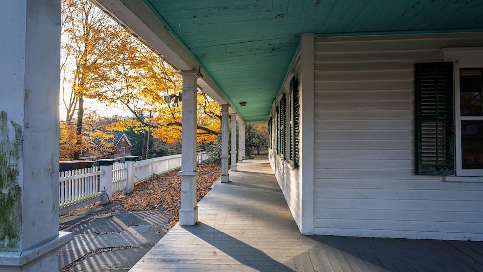 A covered porch with white columns and green roof, overlooking a leaf-covered path and a white fence.