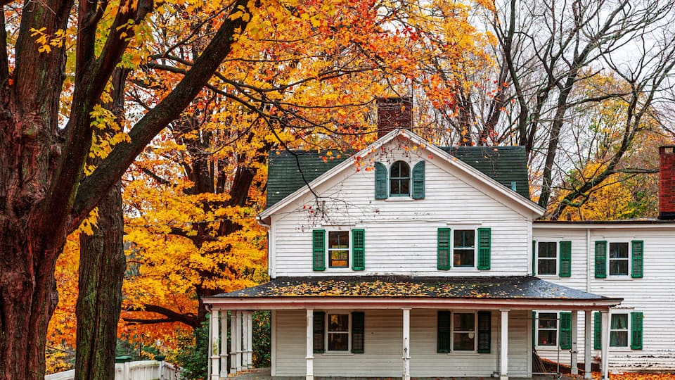 A white house with green shutters and a porch surrounded by autumn trees.